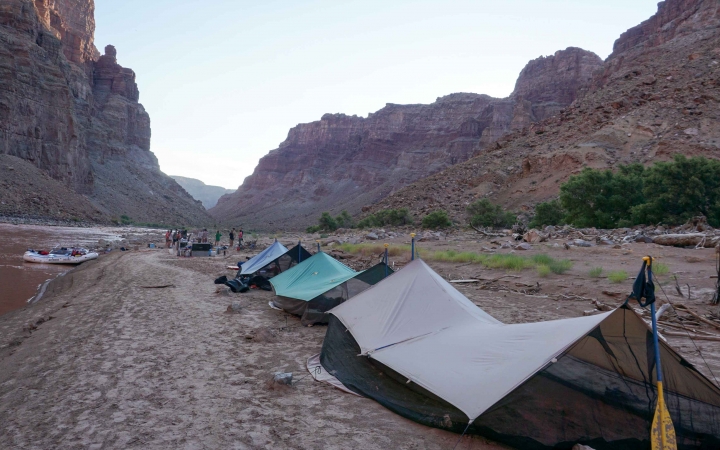 Tarp shelters rest on the bank of a river framed by high canyon walls.